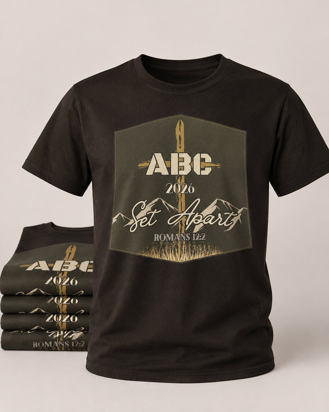 Airsoft Bible Camp 2026 'Set Apart' tee — cross-and-mountains lockup with 'ABC 2026 Set Apart · Romans 12:2' on a brown crew tee, with a stack of olive-green variants beside it
