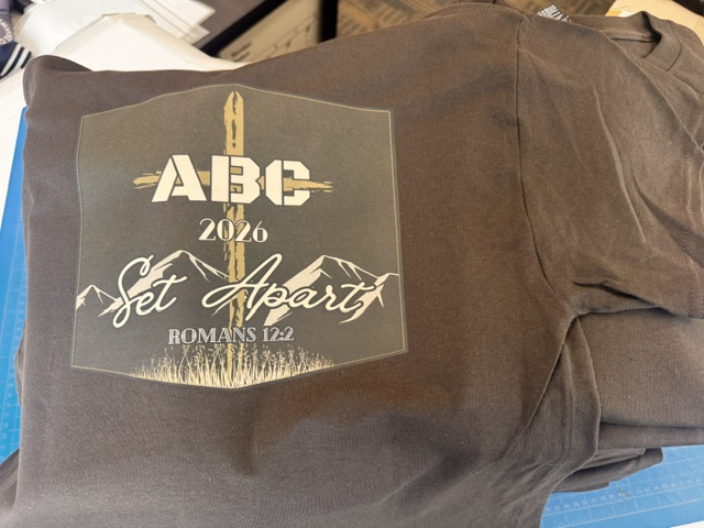 Charcoal grey tee with full-back tone-on-tone print for Airsoft Bible Camp 2026 — Set Apart Romans 12:2 framed by mountains and cross