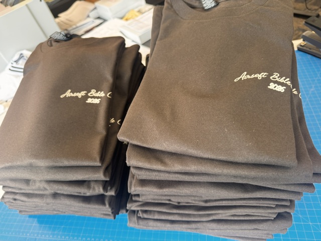 Stack of charcoal camp tees with cream script embroidery for Airsoft Bible Camp 2026