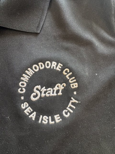 Black staff polo with circular cream-thread embroidery reading 'COMMODORE CLUB · SEA ISLE CITY · Staff'